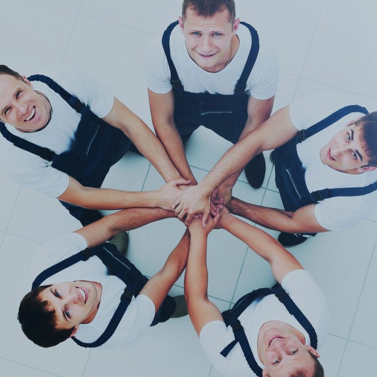 Cinq hommes souriants en salopette, se tenant les mains en cercle.