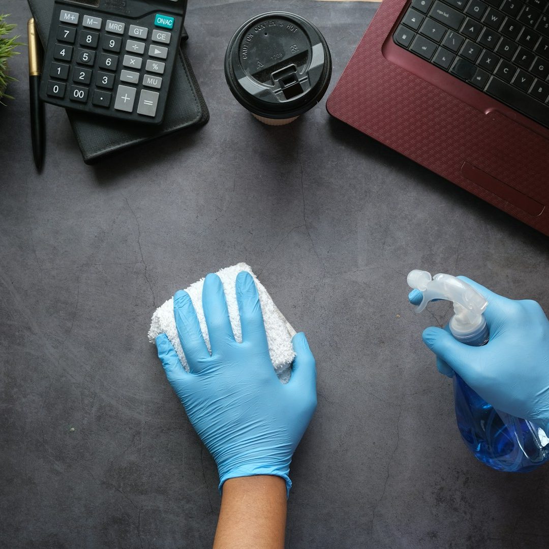 Main d'une personne en gants bleus nettoyant une surface avec un spray et un chiffon.