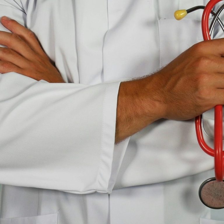 Un médecin en blouse blanche tenant un stéthoscope rouge, avec les bras croisés.